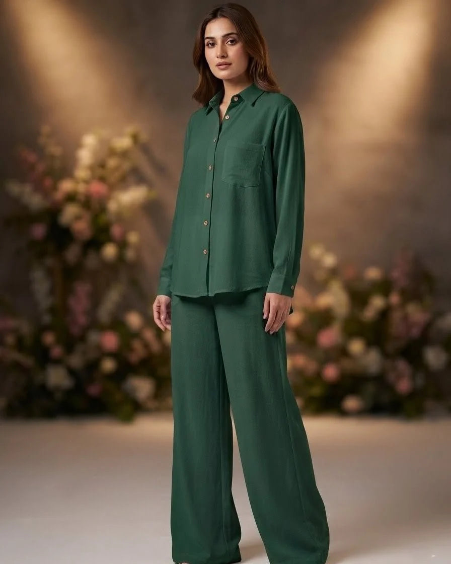 A woman modeling a matching dark green two-piece lounge set consisting of a long-sleeve button-up shirt and wide-leg trousers, set against a warm, softly lit background with blurred floral arrangements.