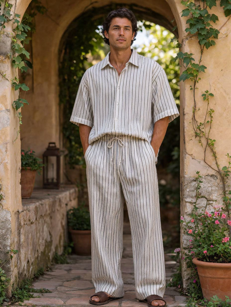 A man wearing a matching short-sleeve button-up shirt and drawstring trousers set in vertical stripes, standing in a stone archway with plants and potted flowers.