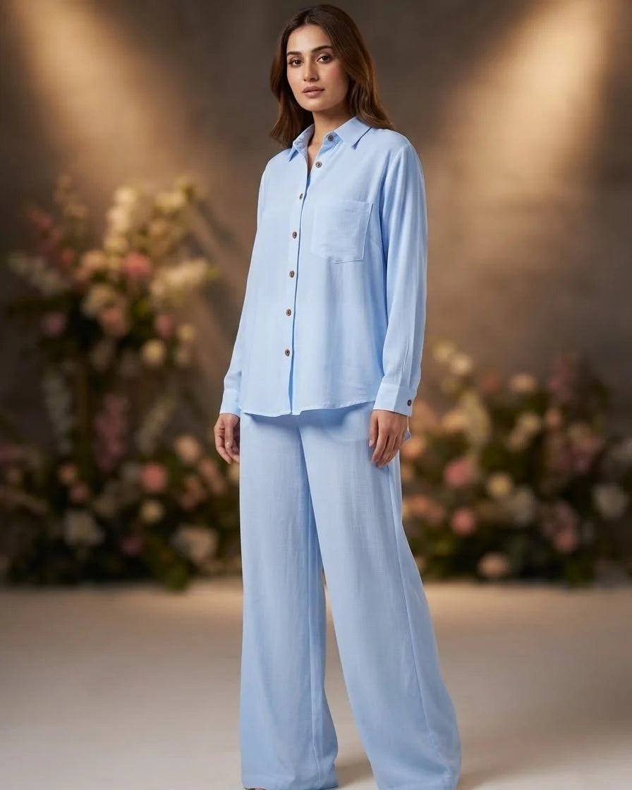 A woman modeling a light blue two-piece pajama set consisting of a long-sleeve button-up shirt and matching wide-leg pants, set against a warm, softly lit background with blurred floral arrangements.