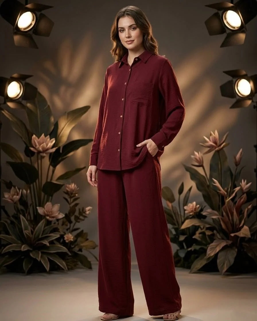A woman modeling a matching burgundy button-up shirt and wide-leg trousers set, standing in a styled studio with warm spotlights and decorative plants.