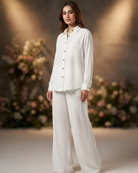 A woman modeling a white long-sleeve button-up shirt and matching wide-leg trousers, standing in a softly lit studio with blurred floral arrangements in the background.
