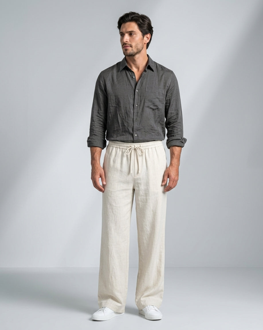 A man modeling a dark gray linen button-up shirt and cream linen drawstring trousers against a light gray studio background.