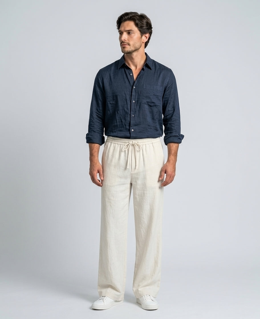 A man modeling a dark navy blue long-sleeve button-up shirt and cream-colored linen drawstring trousers, paired with white sneakers, against a light gray studio background.