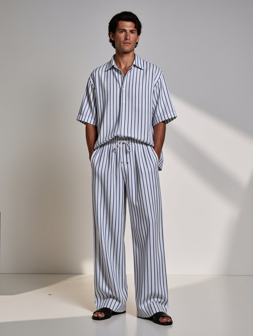 A man wearing a matching short-sleeve button-up shirt and wide-leg trousers set in a blue and white vertical stripe pattern, standing against a minimalist off-white studio background with soft diagonal shadows.